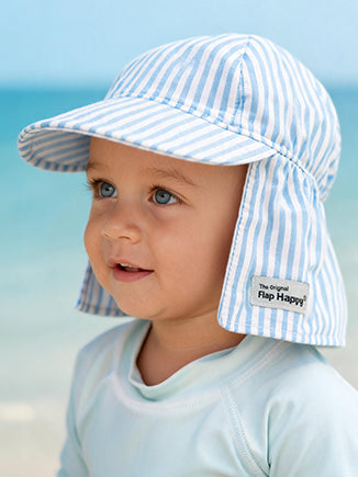 A baby wearing a light blue and white seersucker striped sun hat with a neck flap, photographed on the beach. The hat has a small sewn label that reads 'The Original Flap Happy' visible on the neck flap. 