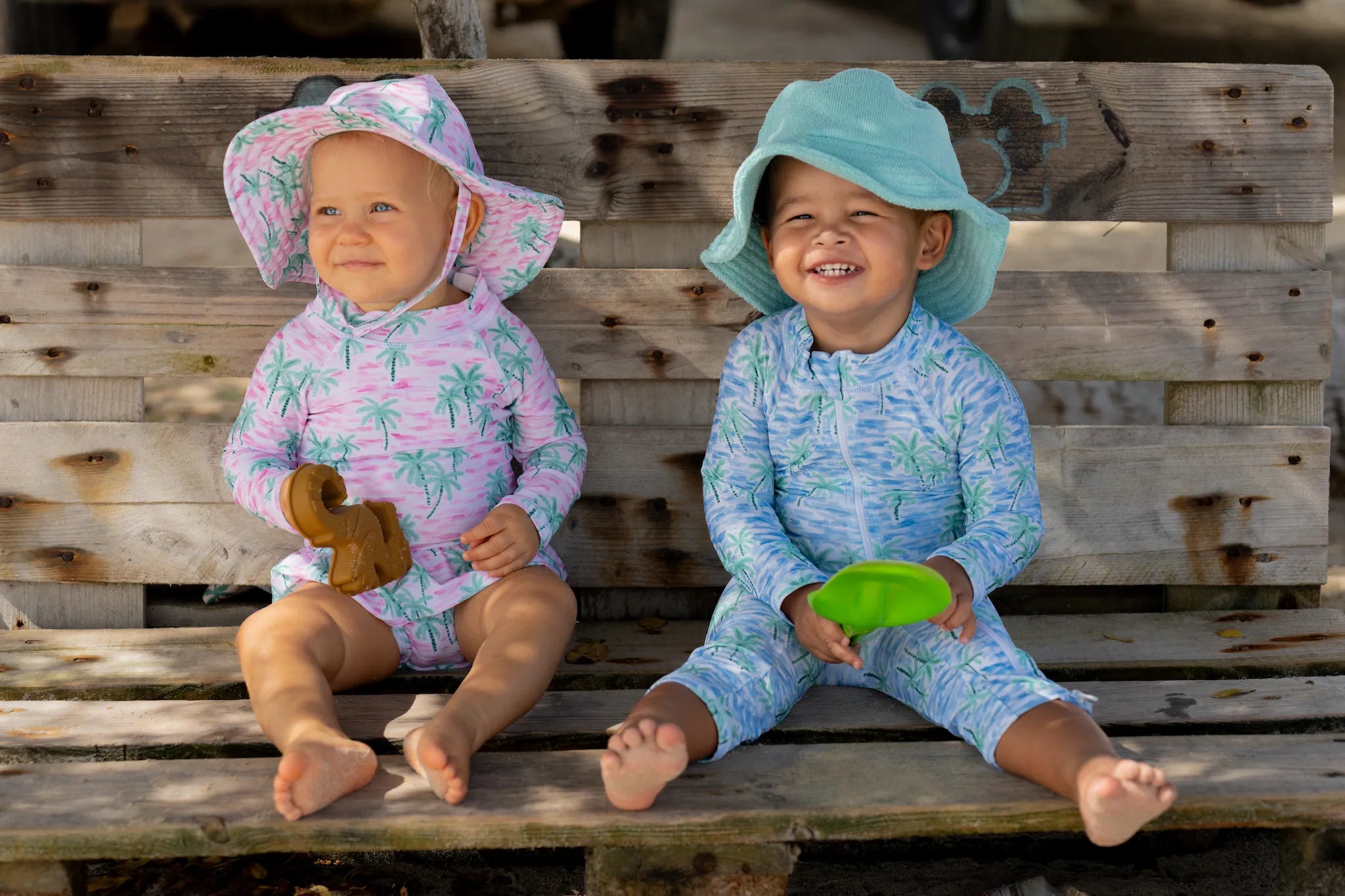 Two children sitting on a wooden bench wearing colorful flap happy rash guard swimsuits and hats.