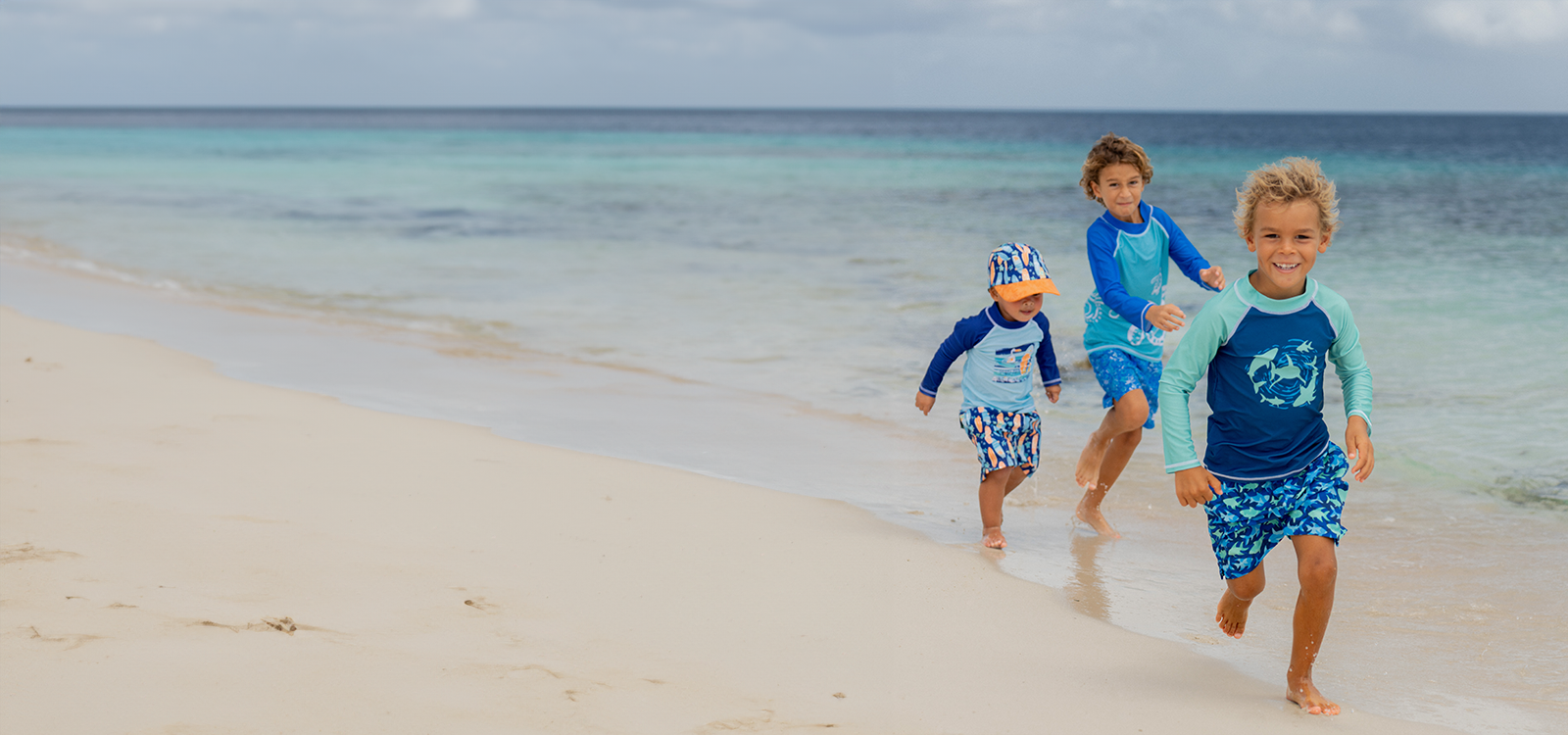 Three children in matching blue and green swimwear running on a sandy beach with ocean in the background.