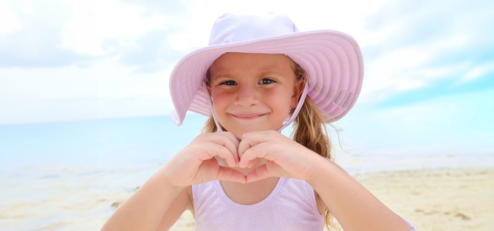 Young girl on a beach wearing a pink hat and making a heart shape with her hands.